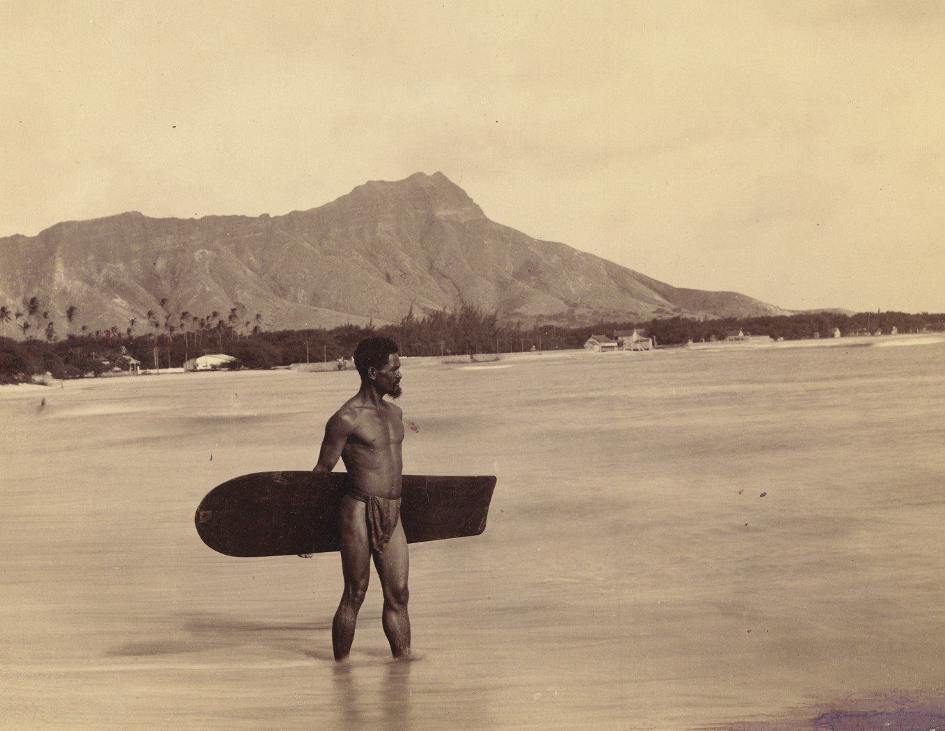 Charles Kauha standing in the shallows at Waikīkī with his alaia surfboard, Diamond Head behind him — Frank Davey photograph, April 1898